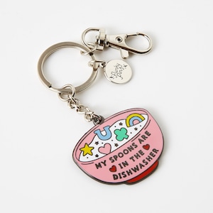 Puede incluir: Un llavero de esmalte rosa con un dise&ntilde;o de cuenco. El cuenco presenta un arco&iacute;ris, estrellas, corazones y el texto "My spoons are in the dishwasher". El llavero tiene un llavero plateado, un cierre y un peque&ntilde;o dije con el texto "Punky Pins."