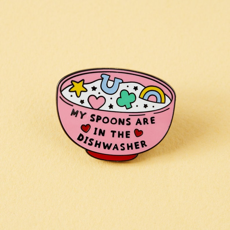 May include: Pink enamel pin shaped like a bowl of cereal with the text "My spoons are in the dishwasher" and colorful cartoon shapes inside.