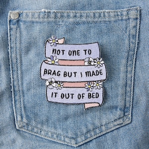 Könnte beinhalten: Ein gestickter Aufnäher in Lila und Rosa mit dem Text "Not one to brag but I made it out of bed" und weißen Gänseblümchen um den Text herum.