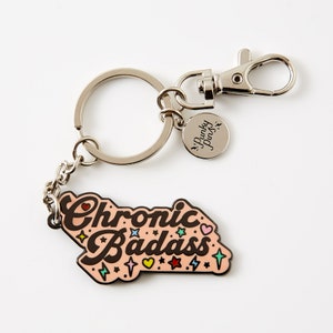 Puede incluir: Un llavero de metal rosa con el texto "Chronic Badass" en letras negras con estrellas y corazones de colores. El llavero tiene un anillo de metal plateado y un dije plateado con el texto "Punky Pins".