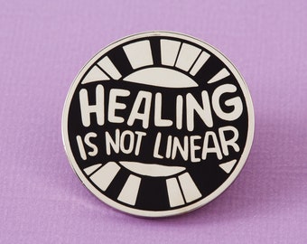 May include: Black and white enamel pin with the text "Healing is not linear" in a white circle with a black background. The circle has a black and white striped design around the edge.