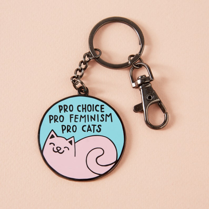 Puede incluir: Un llavero redondo con fondo azul claro y las palabras "PRO CHOICE PRO FEMINISM PRO CATS" en texto negro. Una ilustraci&oacute;n de gato rosa est&aacute; en el centro. El llavero tiene un cierre y un llavero plateados.