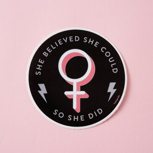 Può includere: Un adesivo bianco e nero con un simbolo femminile rosa e il testo "Credeva di potercela fare, quindi l'ha fatto".