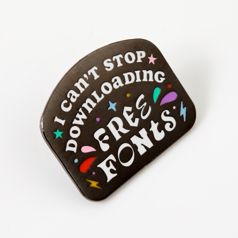 May include: Black enamel pin with white text that reads "I can't stop downloading free fonts". The pin is decorated with colorful stars, lightning bolts, and teardrops.