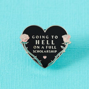 May include: Black heart-shaped enamel pin with a silver outline. The pin features a rose on each side and the text "Going to Hell on a Full Scholarship".