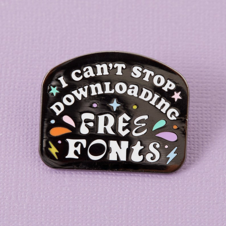 May include: Black enamel pin with white text that reads "I can't stop downloading free fonts". The pin is decorated with colorful stars, lightning bolts, and swirls.