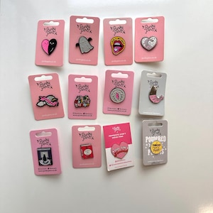 Pode incluir: Doze pinos de esmalte em cartões cor-de-rosa com o texto "Punky Pins" em cada cartão. Os pinos apresentam vários desenhos, incluindo um fantasma, um coração, um livro, um pássaro e um símbolo feminista.