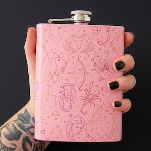May include: A pink hip flask with a silver cap and hinge. The flask features gold line drawings of zodiac symbols, such as a crab, lion, and scorpion. The flask is set against a black backdrop.