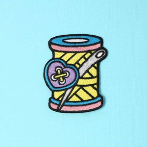 May include: An embroidered patch depicting a spool of thread with a needle and a purple heart-shaped button, in yellow and pink.