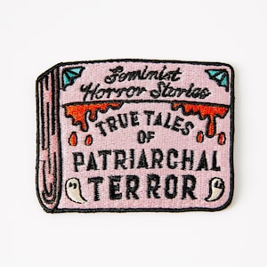 Könnte beinhalten: Ein gestickter Aufnäher in Form eines Buches mit dem Text "Feminist Horror Stories True Tales of Patriarchal Terror" und zwei Geisterfiguren.