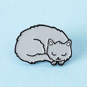 Könnte beinhalten: Eine graue Emaille-Anstecknadel mit einem schlafenden, zusammengerollten Kätzchen. Die Katze ist schwarz umrandet und hat ein einfaches, cartoonartiges Design.