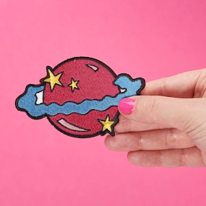Puede incluir: Parche bordado que representa un planeta rojo con formas azules en forma de nube y estrellas amarillas. El parche tiene un borde cosido y se sostiene sobre un fondo rosa. El diseño es caprichoso y colorido, adecuado para adornar ropa o accesorios.