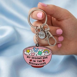 Puede incluir: Un llavero con un dise&ntilde;o de cuenco rosa. El cuenco tiene un borde rojo y est&aacute; lleno de varias formas coloridas y el texto "MY SPOONS ARE IN THE DISHWASHER". El llavero incluye un llavero plateado, un mosquet&oacute;n y un peque&ntilde;o dije con el texto "Punky Pins".