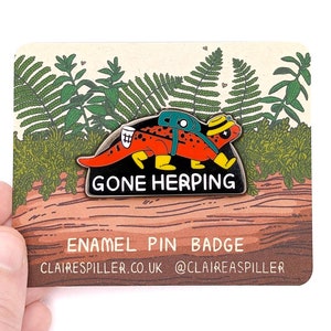 May include: A red and orange enamel pin badge featuring a cartoon salamander with a backpack and the text "Gone Herping". The pin is set against a background of green ferns and brown wood.