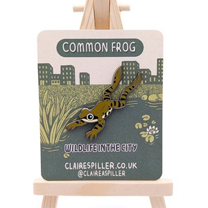 Op de afbeelding: Een speld met een groen en bruin kikkerontwerp met zwarte strepen, gepresenteerd op een kaart. De kaart leest "COMMON FROG" en "WILDLIFE IN THE CITY". De achtergrond is groen, met een vijverlandschap en een skyline.
