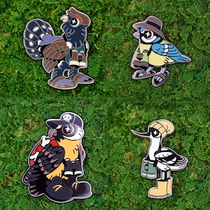 Könnte beinhalten: Vier Emaille-Anstecknadeln mit anthropomorphen Vögeln. Die Anstecknadeln zeigen einen Hahn, eine Blauracke, eine Eule und einen Schnepfenvogel, jeweils mit einzigartigen Outfits und Accessoires. Die Anstecknadeln sind vor einem grünen, strukturierten Hintergrund angeordnet.