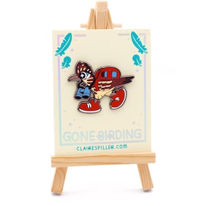 May include: A colorful enamel pin featuring a cartoon bird with binoculars and red sneakers, displayed on a small wooden easel. The pin is on a card that says "Gone Birding" and "clairespiller.com".