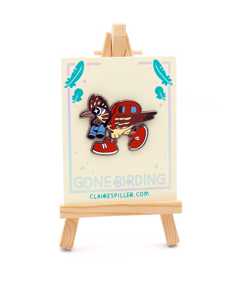 May include: A colorful enamel pin featuring a cartoon bird with binoculars and red sneakers, displayed on a small wooden easel. The pin is on a card that says "Gone Birding" and "clairespiller.com".
