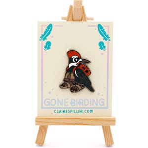 May include: A pin featuring a cartoon woodpecker in a red and black outfit, displayed on a small wooden easel. The pin is on a cream-colored card with the text "GONE BIRDING" and "clairespiller.com". Teal feather and jellyfish graphics are on the card.