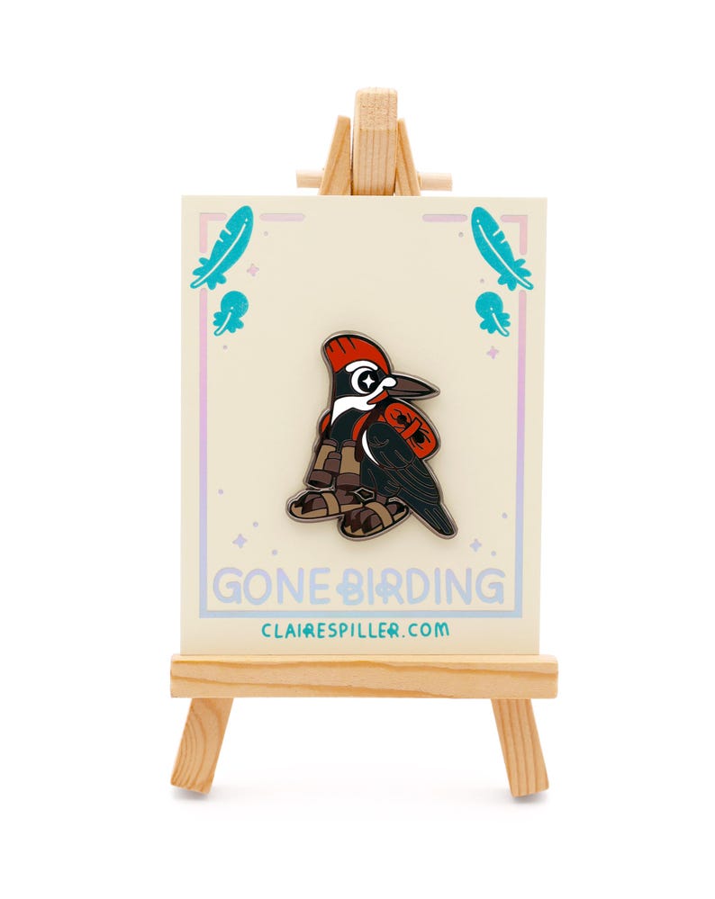 May include: A pin featuring a cartoon woodpecker in a red and black outfit, displayed on a small wooden easel. The pin is on a cream-colored card with the text "GONE BIRDING" and "clairespiller.com". Teal feather and jellyfish graphics are on the card.