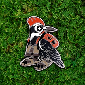 May include: A detailed enamel pin featuring a stylized woodpecker design. The pin showcases a black body, red head and accents, and a white eye with a star. The woodpecker is set against a backdrop of green moss.