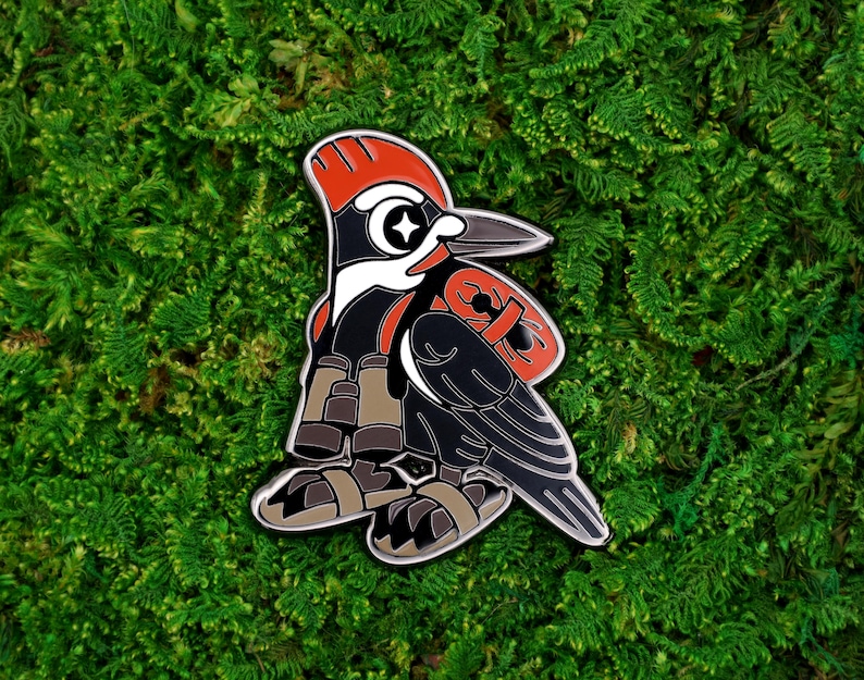 May include: A detailed enamel pin featuring a stylized woodpecker design. The pin showcases a black body, red head and accents, and a white eye with a star. The woodpecker is set against a backdrop of green moss.