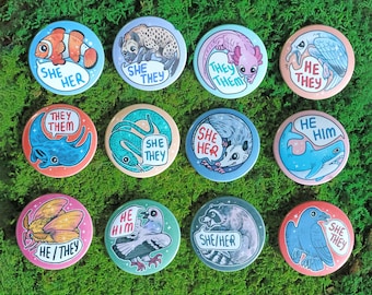 May include: A set of twelve circular pin-back buttons featuring various animal illustrations and pronoun text. Animals include a clownfish, hyena, axolotl, bird, stingray, snake, opossum, shark, moth, pigeon, raccoon, and crow with pronouns like she/her, they/them, he/him, and he/they.