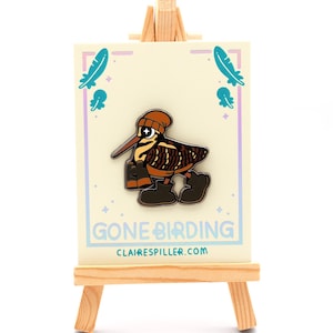 May include: A whimsical enamel pin featuring a cartoon bird wearing a beanie and boots. The pin is on a card with the text "GONE BIRDING" and the website clairespiller.com. The card is displayed on a small wooden easel.