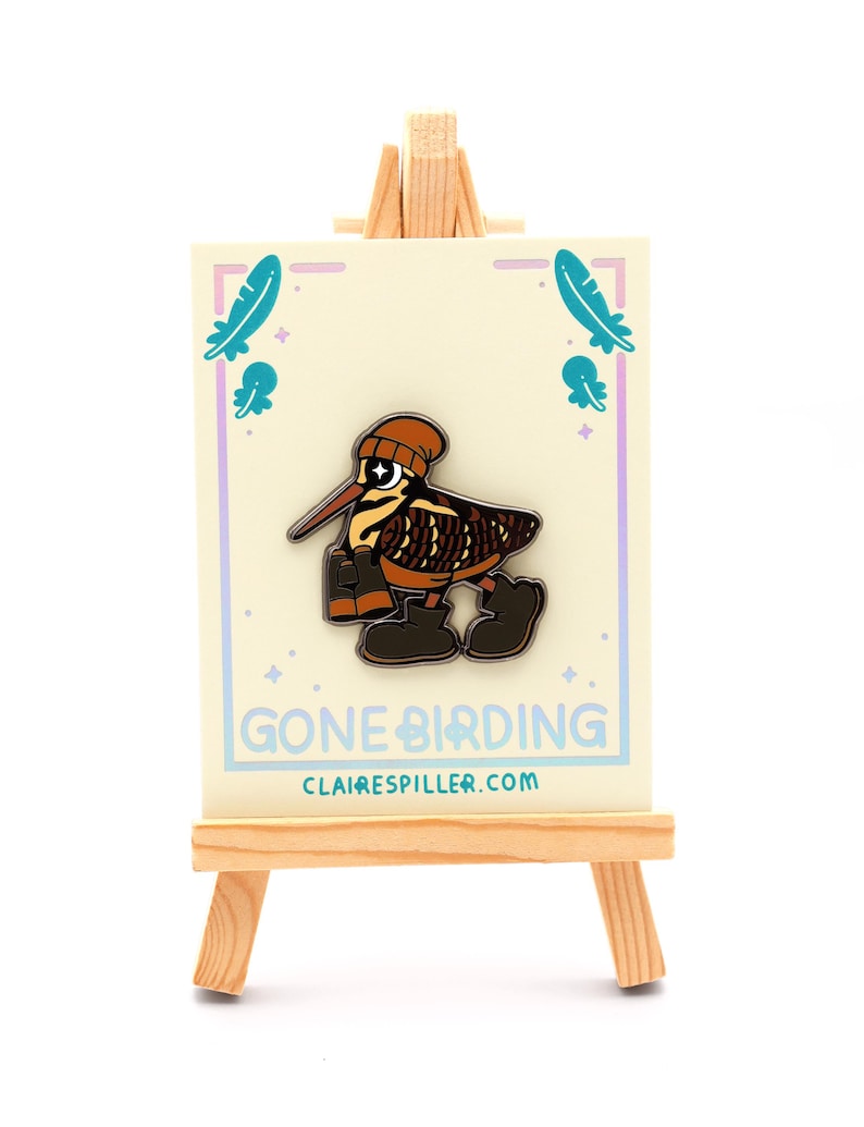 May include: A whimsical enamel pin featuring a cartoon bird wearing a beanie and boots. The pin is on a card with the text "GONE BIRDING" and the website clairespiller.com. The card is displayed on a small wooden easel.