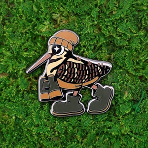 May include: A whimsical enamel pin featuring a cartoon bird with a brown beanie, sunglasses, and boots. The bird has a long beak and is holding binoculars. The pin is set against a green moss background.