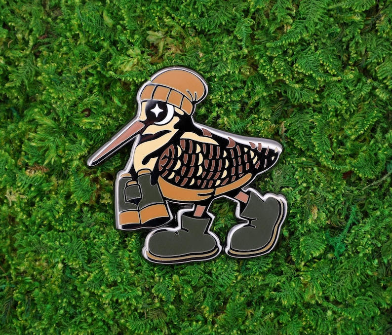 May include: A whimsical enamel pin featuring a cartoon bird with a brown beanie, sunglasses, and boots. The bird has a long beak and is holding binoculars. The pin is set against a green moss background.