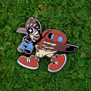 May include: A colorful enamel pin featuring a cartoon roadrunner character with binoculars. The pin has a brown, tan, and red color scheme, with blue accents on the binoculars and backpack. The roadrunner is wearing red shoes with white soles.
