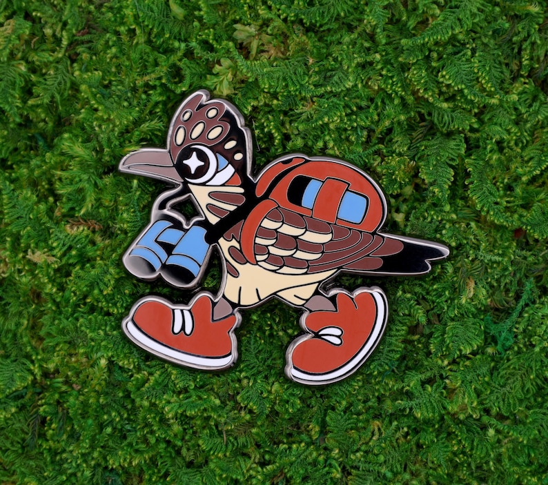 May include: A colorful enamel pin featuring a cartoon roadrunner character with binoculars. The pin has a brown, tan, and red color scheme, with blue accents on the binoculars and backpack. The roadrunner is wearing red shoes with white soles.