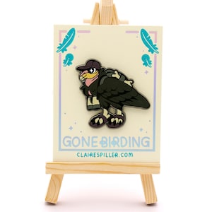 May include: A pin featuring a cartoon vulture wearing a cap and backpack, with the text "GONE BIRDING" and the website clairespiller.com. The pin is displayed on a small wooden easel against a white background.