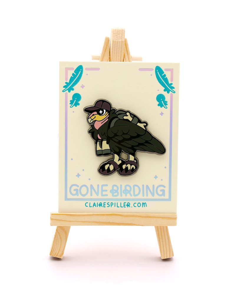 May include: A pin featuring a cartoon vulture wearing a cap and backpack, with the text "GONE BIRDING" and the website clairespiller.com. The pin is displayed on a small wooden easel against a white background.