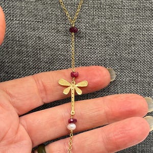 Op de afbeelding: Een goudkleurige ketting met een libelle hanger. De ketting heeft een fijne ketting met rode en witte kralen. De libelle hanger is een gestileerd silhouet. De ketting wordt gepresenteerd tegen een grijze achtergrond.