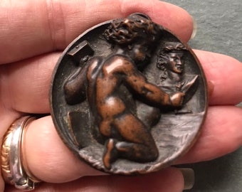 Può includere: Un ciondolo circolare in bronzo con una scultura a rilievo di un cherubino nudo che tiene uno scalpello e un libro. Il cherubino è inginocchiato e guarda il libro. C'è una scultura a rilievo più piccola di una testa di persona sullo sfondo.