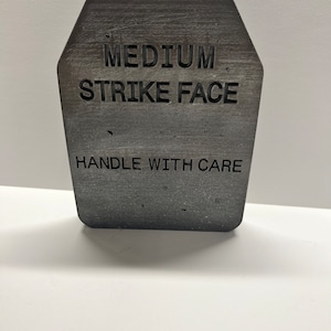 Puede incluir: Una placa de impacto rectangular gris con el texto "MEDIUM STRIKE FACE" y "HANDLE WITH CARE" grabado en ella.