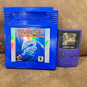 Könnte beinhalten: Eine blaue Game Boy Color-Cartridge für das Spiel Yu-Gi-Oh! Die Cartridge befindet sich neben einer violetten Game Boy Color-Handheld-Konsole. Der Text auf der Cartridge lautet "Yu-Gi-Oh!" und "KONAMI".