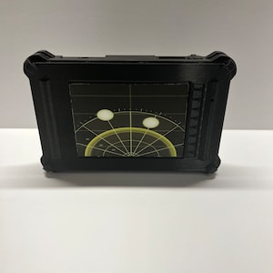 Op de afbeelding: Een zwart, rechthoekig apparaat met een radar-achtig display. Het scherm toont een geel en zwart grafisch ontwerp met een cirkelvormig patroon en twee witte cirkels. Het apparaat heeft een getextureerde lijst, tegen een witte achtergrond.