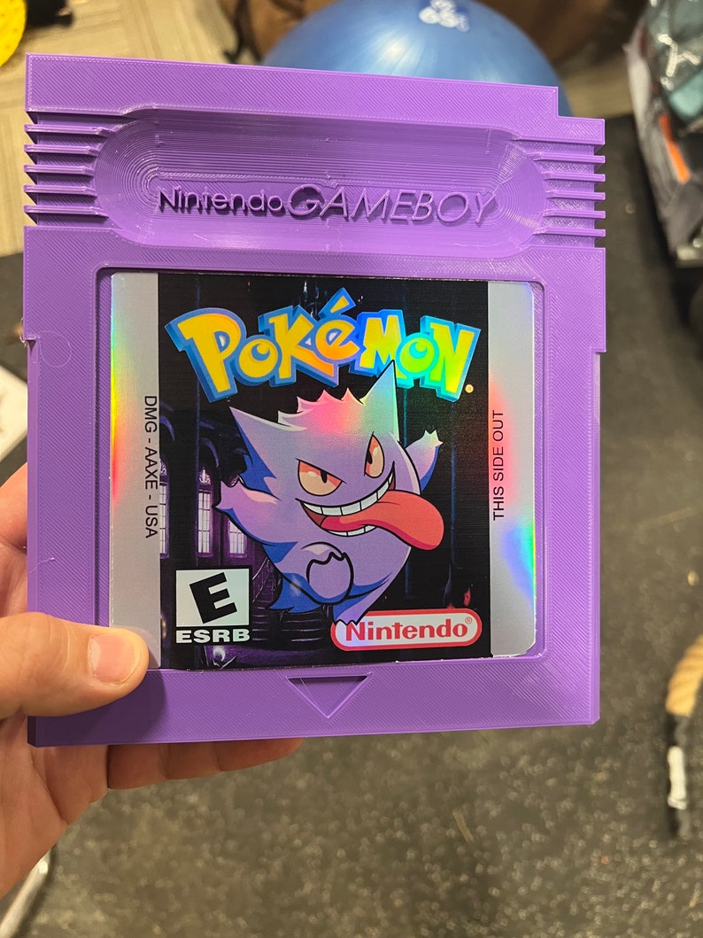 May include: Purple Game Boy Color cartridge with a holographic label featuring a purple and white Pokemon character with a large tongue. The label reads "Pokemon" and "Nintendo". The ESRB rating "E" is visible on the label.