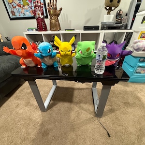 May include: A black gaming desk with silver legs supports a collection of colorful Pokémon plush toys, including Charmander, Squirtle, Pikachu, Bulbasaur, and Gengar. A water bottle sits among the toys.
