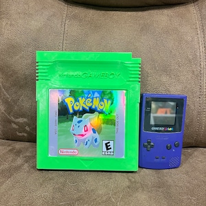 Puede incluir: Cartucho verde de Game Boy Color con una imagen holográfica de un personaje Pokemon azul en la parte delantera. El cartucho está etiquetado como "Pokemon" y "Nintendo Game Boy". El cartucho está al lado de una consola Game Boy Color morada.