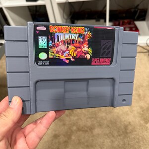 Könnte beinhalten: Eine graue Super Nintendo-Spielkassette für Donkey Kong Country. Das Etikett zeigt den Titel und die Charaktere des Spiels sowie die Nintendo- und ESRB-Logos. Die Kassette wird in einer Hand gehalten.