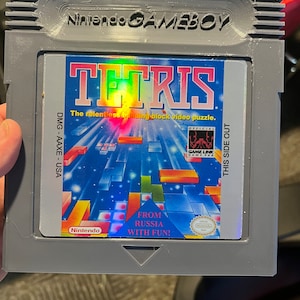 May include: A gray Game Boy cartridge with a colorful label that reads "TETRIS The relentless building block video puzzle." The label also features the text "FROM RUSSIA WITH FUN!" and a Nintendo seal.
