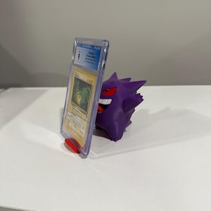 Op de afbeelding: Een paarse 3D-geprinte Gengar-figuur die een beoordeelde Pokémon-ruilkaart vasthoudt. De kaart zit in een doorzichtige plastic hoes met een CGC Universal Grade-label. De Gengar-figuur heeft rode ogen en een ondeugende grijns.