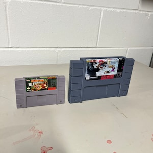 May include: Two Super Nintendo Entertainment System game cartridges. The cartridge on the left is Donkey Kong Country, the cartridge on the right is Super Mario World.