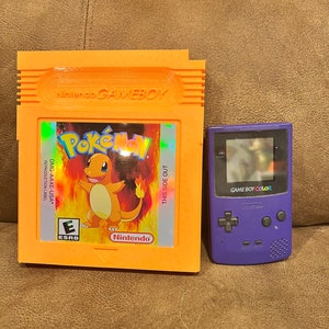 Könnte beinhalten: Eine violette Game Boy Color Handheld-Spielkonsole neben einer Game Boy-Cartridge mit einem Pokemon-Bild. Die Cartridge ist orange und hat den Text "Nintendo GAME BOY" und "Pokemon" darauf. Der Text "THIS SIDE OUT" befindet sich auf der rechten Seite der Cartridge. Die Game Boy Color hat den Text "GAME BOY COLOR" darauf.