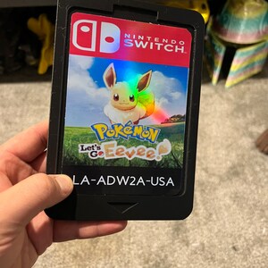 May include: A black Nintendo Switch game cartridge case with a Pokemon Let's Go Eevee game card inside. The game card features a cartoon Eevee on a green grassy field with a blue sky and white clouds. The text "Pokemon Let's Go Eevee!" is printed on the game card. The case has the Nintendo Switch logo and the text "Nintendo Switch" printed on the front. The case also has the text "LA-ADW2A-USA" printed on the bottom.