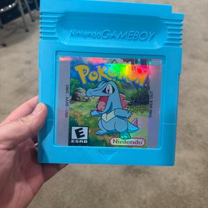 Puede incluir: Un cartucho azul de Game Boy Color con una etiqueta holográfica que muestra el logotipo de Pokemon y un personaje Pokemon azul. La etiqueta también incluye el texto "Nintendo" y "ESRB E".